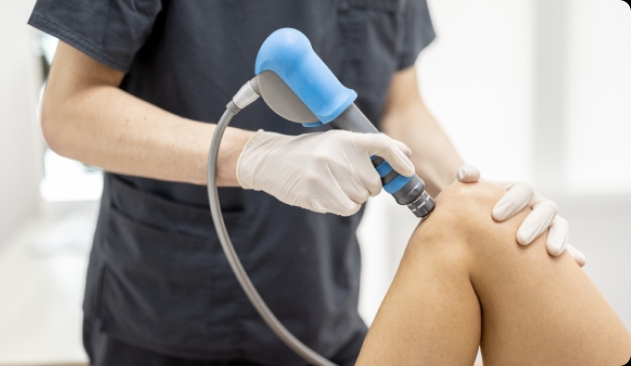 Joelho sendo avaliado a partir da terapia por onda de choque. O aparelho com laterais circulares está sendo segurado por um profissional com luvas. A imagem mostra apenas o joelho, o  aparelho e as mãos do especialista.